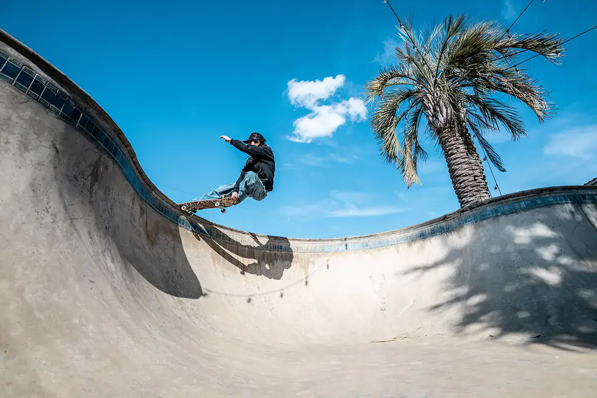 Urban sports photography of skateboarder performing trick in concrete bowl under blue sky and palm tree.