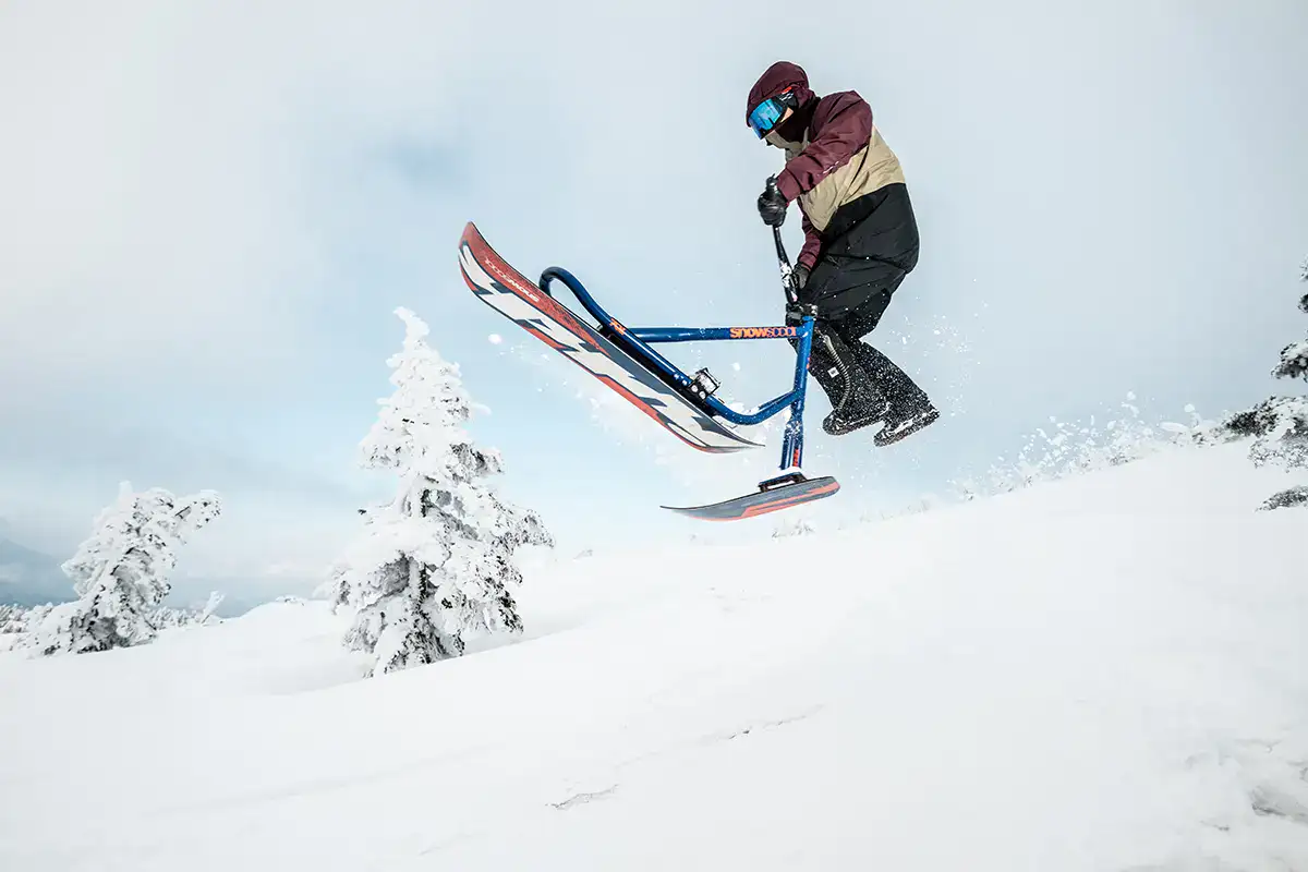 Urban sports photography showcasing rider performing snowscoot jump on snowy mountain with dramatic winter landscape