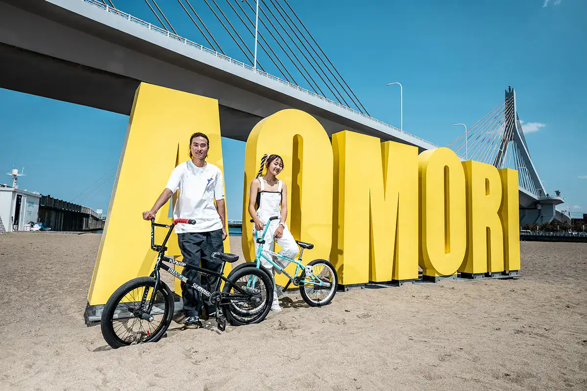 Urban sports photography of BMX riders posing at Aomori beach with landmark sign and bridge backdrop.