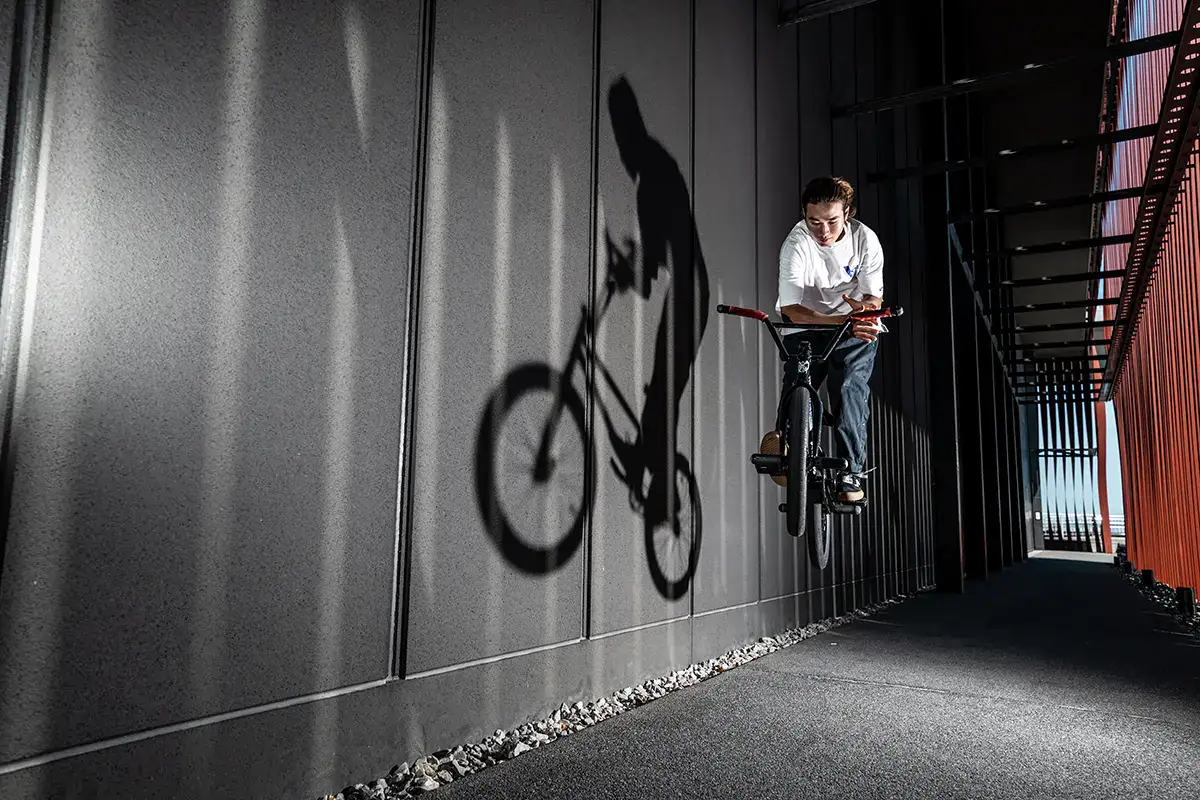 BMX rider jumping beside Wa Rasse building, casting dramatic shadow across the wall in sunlight.