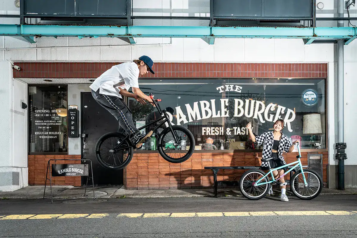 BMX rider performs jump outside Ramble Burger shop while another rider watches and smiles.