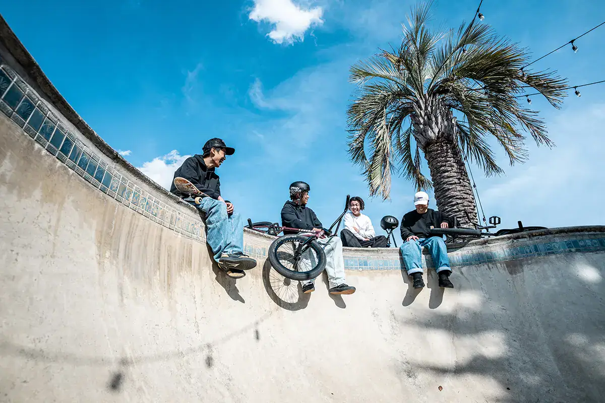 Group of BMX riders relaxing with bikes at skatepark bowl under blue sky and palm tree.