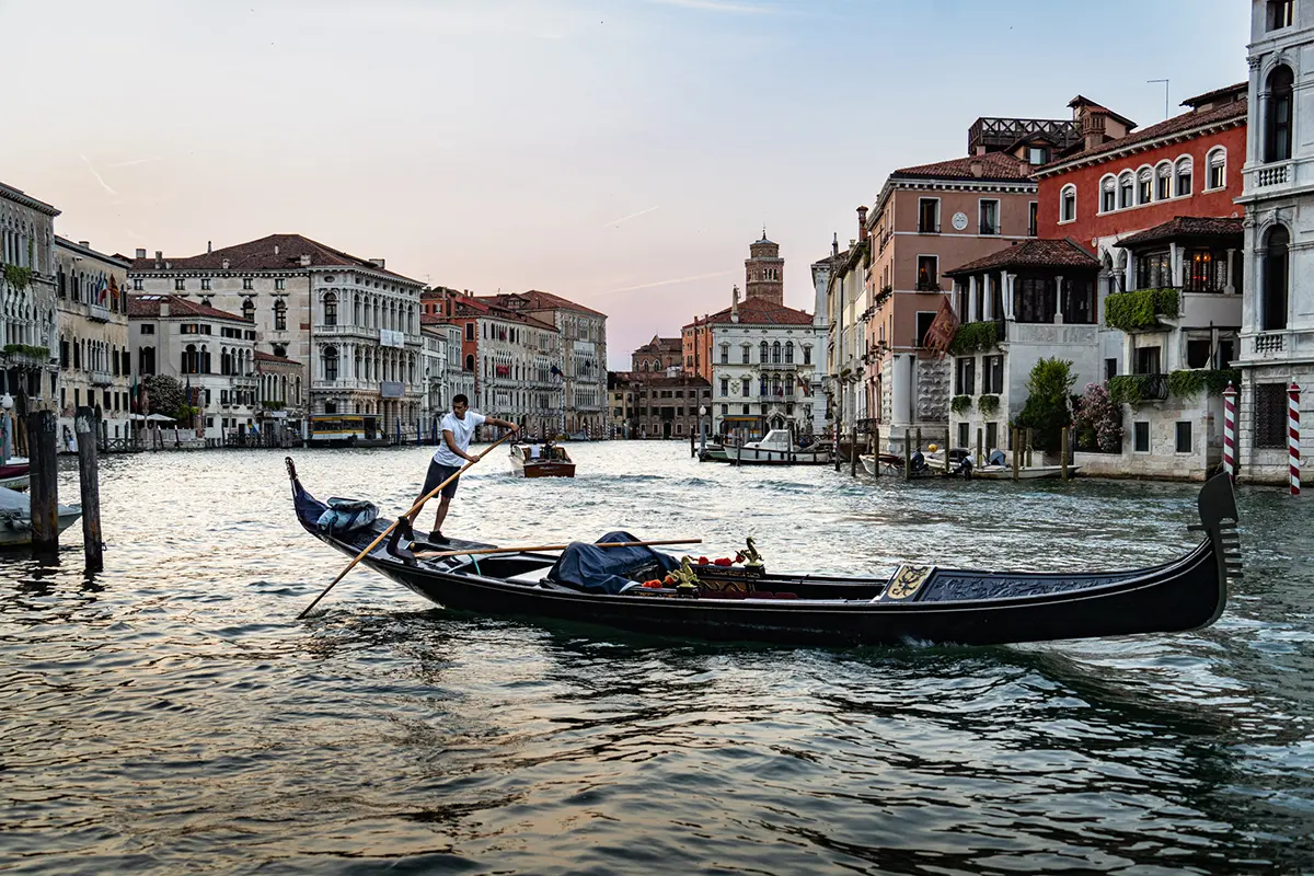 A gondolier rows a traditional gondola along the Grand Canal in Venice at sunset, surrounded by historic buildings.