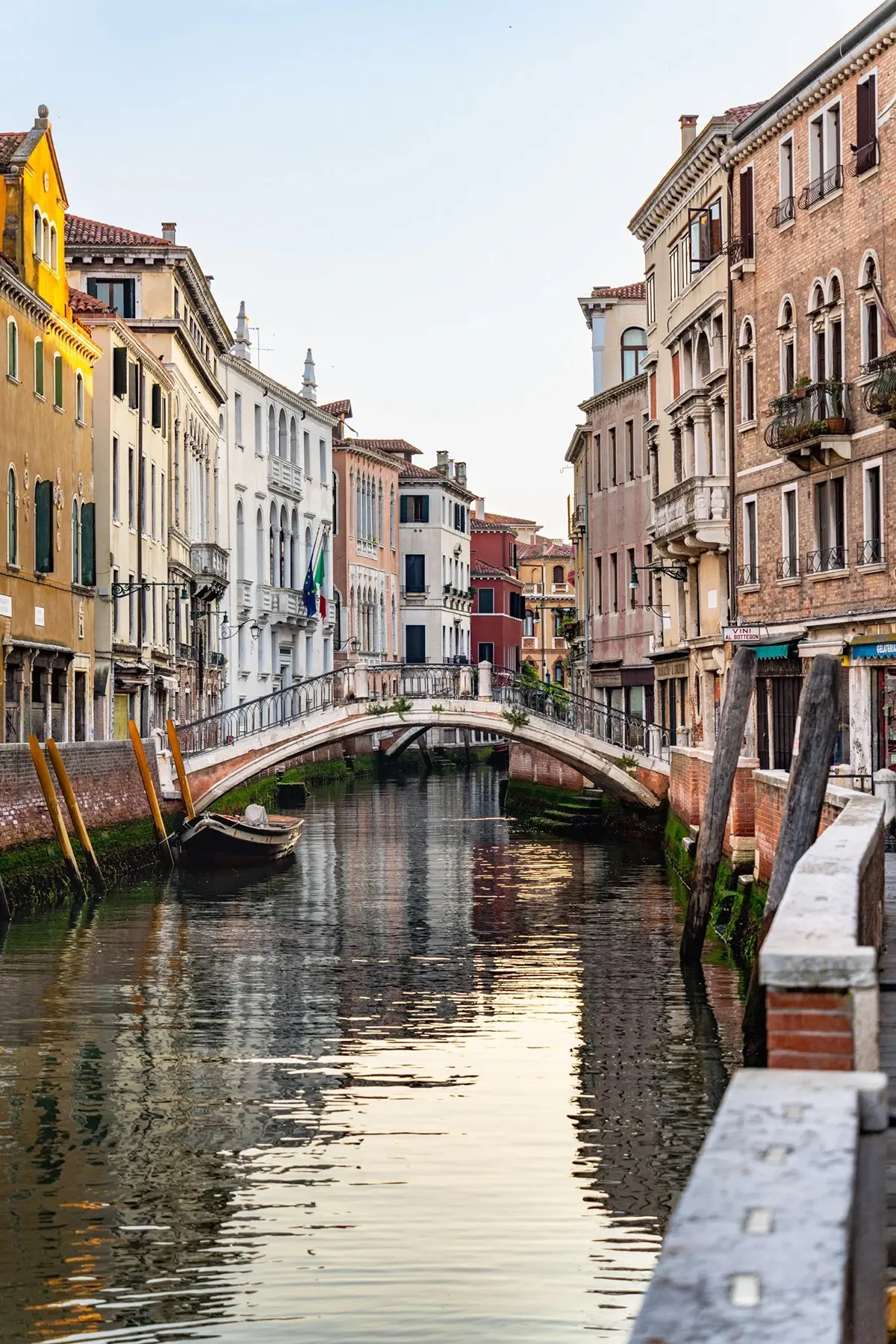A stone footbridge spans a quiet Venetian canal at sunset, with reflections of historic buildings in the water.