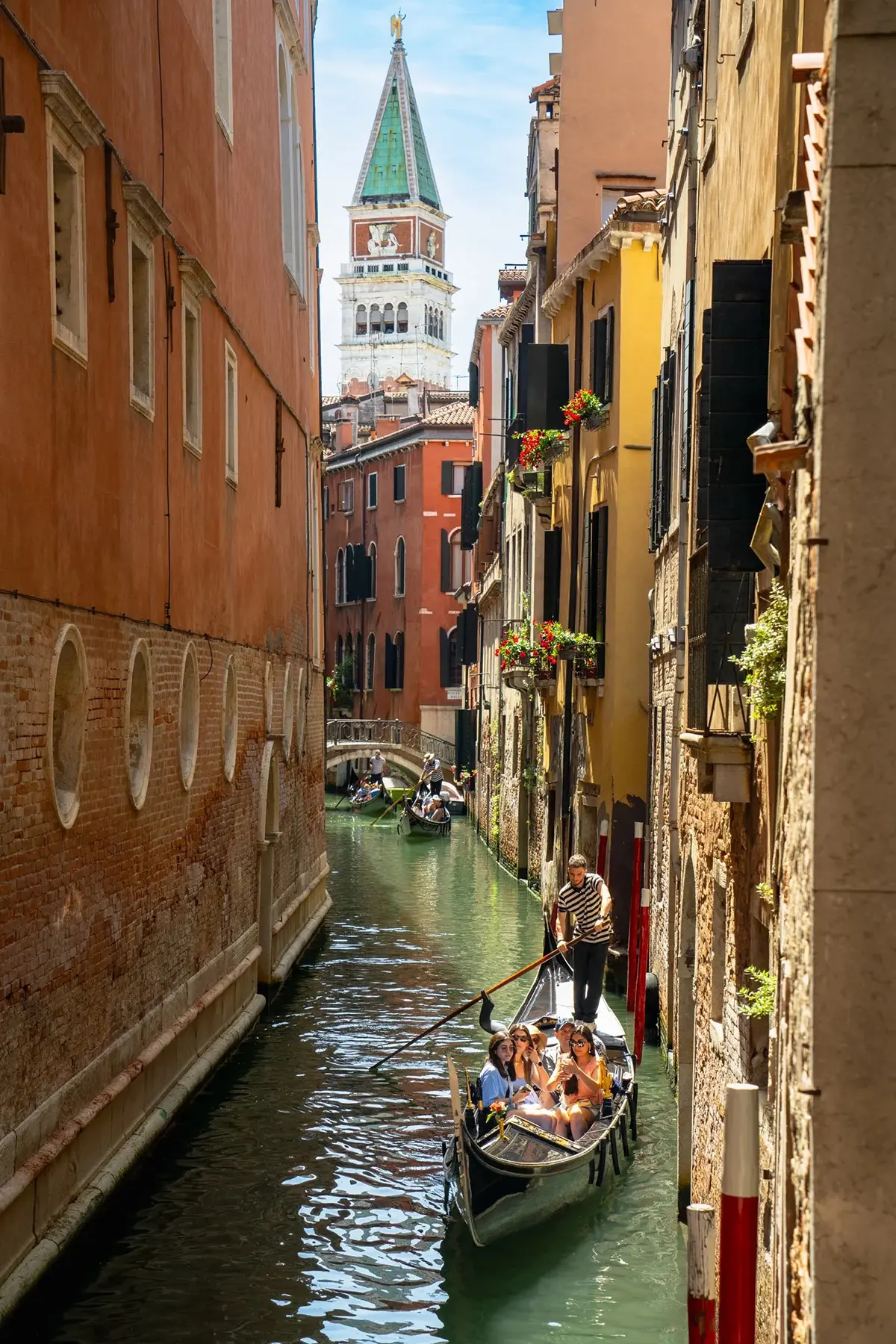 A gondolier rows tourists through a narrow Venetian canal with the Campanile di San Marco tower rising in the background.