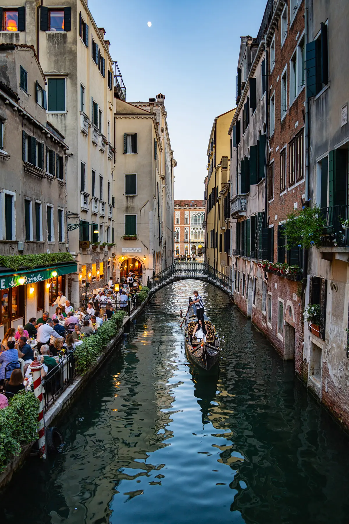 A gondolier rows past a lively canal-side restaurant in Venice at dusk, with diners enjoying the evening atmosphere.