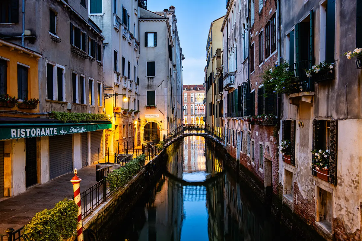 Evening view of a quiet Venetian canal with warm lights reflecting on the water and a small footbridge in the distance.