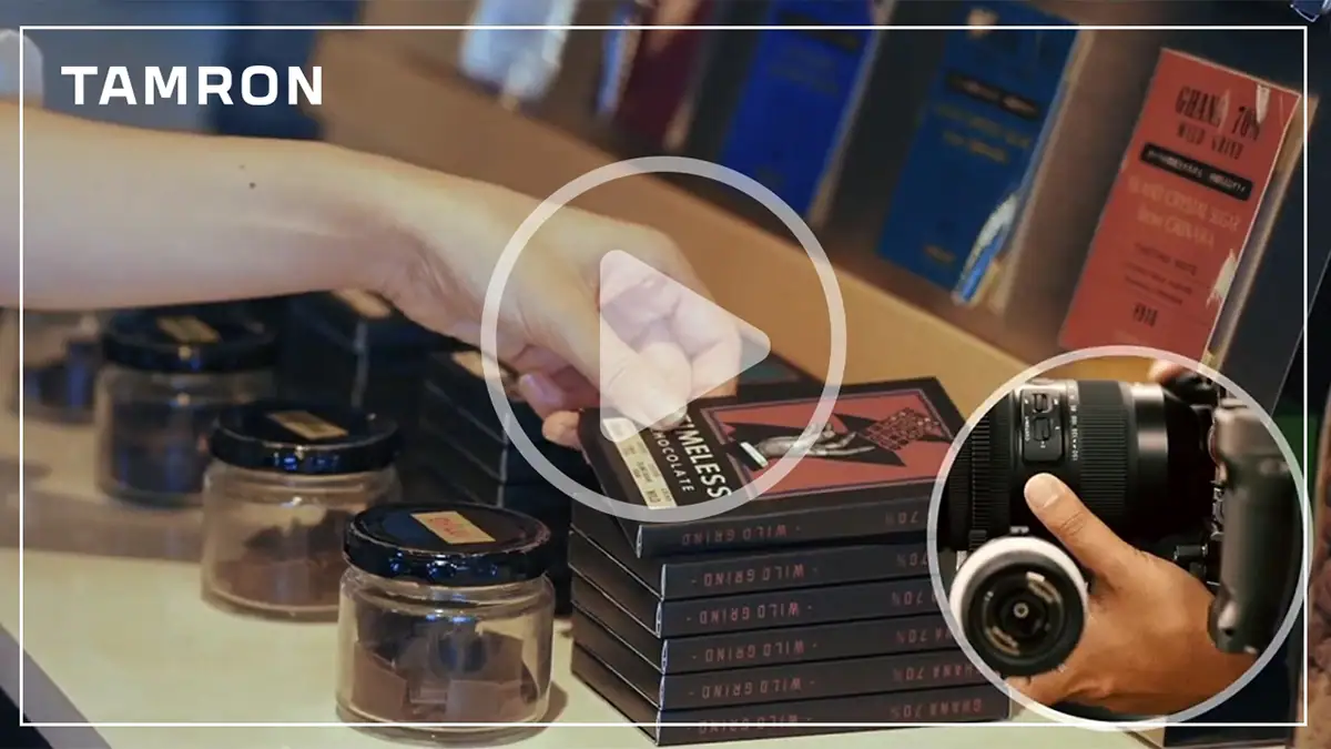 Thumbnail for a video demonstrating A-B Focus functionality using the TAMRON Lens Utility, showing a shopper&rsquo;s hand reaching for chocolate and a close-up of a camera with a TAMRON lens.