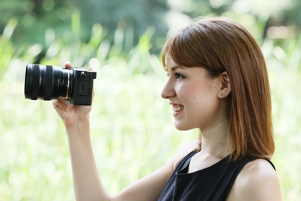 Smiling woman filming with a compact mirrorless camera and zoom lens outdoors, holding it in selfie or vlogging position with a blurred green background.