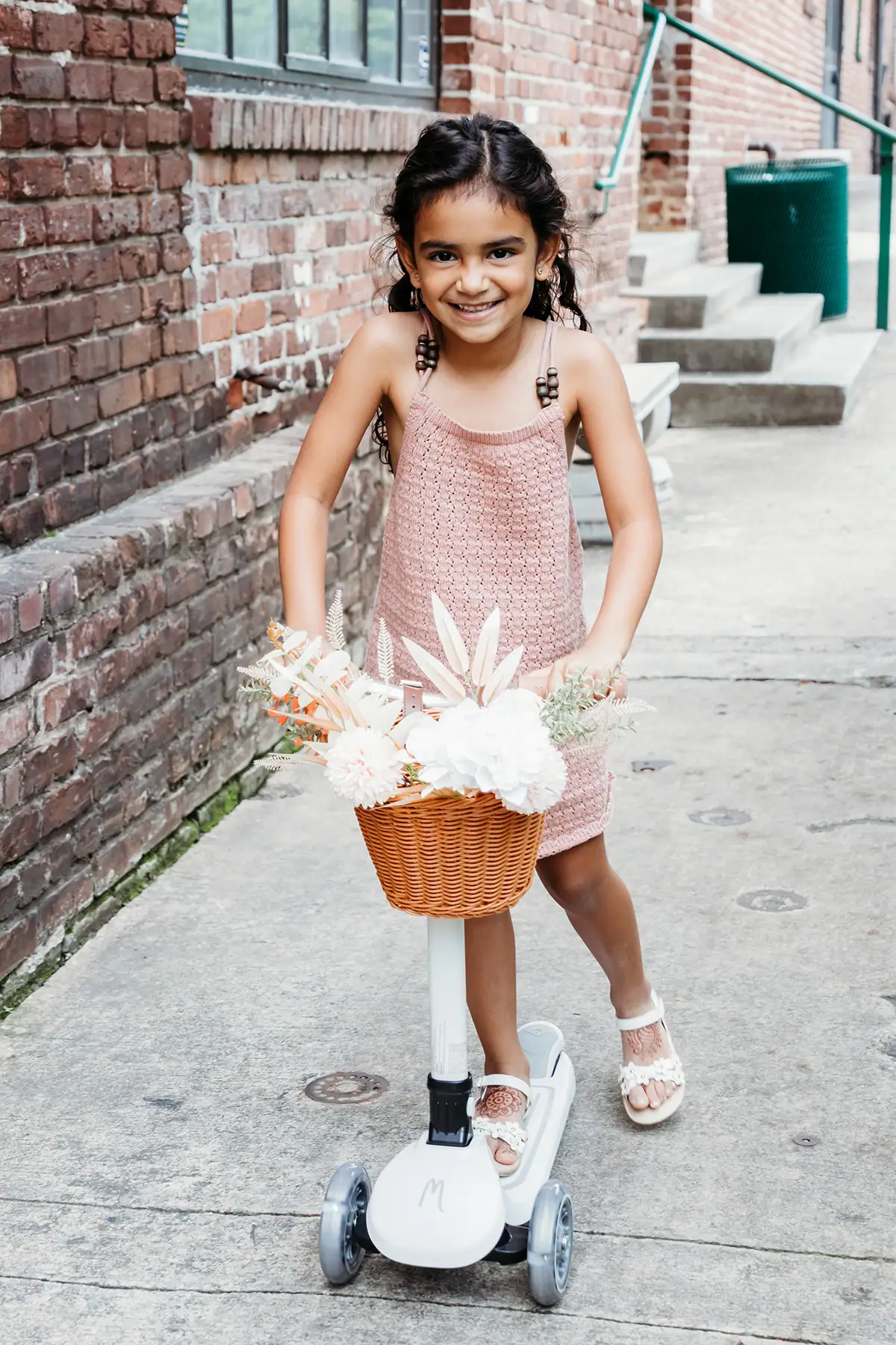 Smiling girl riding a scooter with flowers in the basket during a family photography session outdoors.