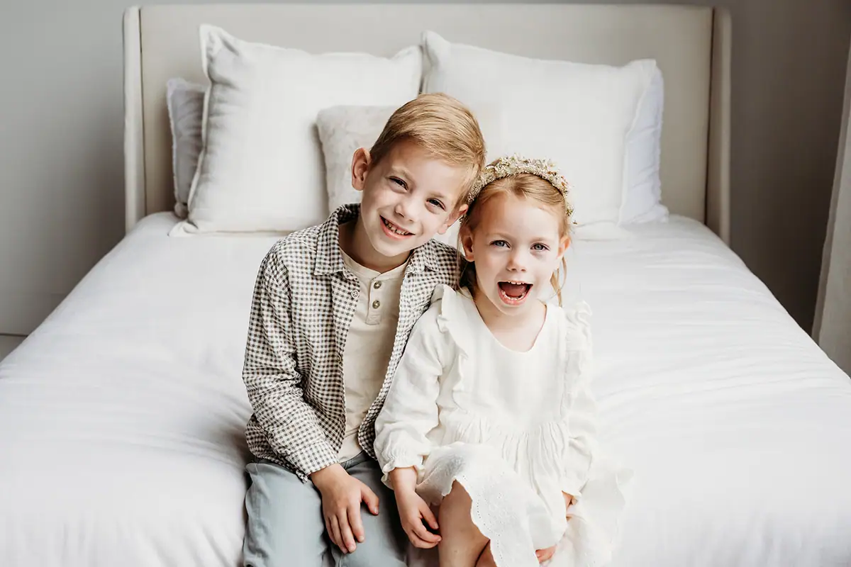 Brother and sister sitting on a bed by the window smiling during an indoor family photography session.