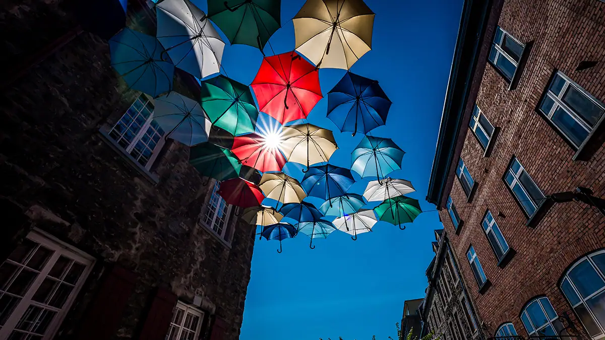 A vibrant street scene with colorful umbrellas suspended overhead, forming a canopy against a clear blue sky, with the sun's rays visible through them and framed by old, brick buildings on either side, featuring multiple windows.