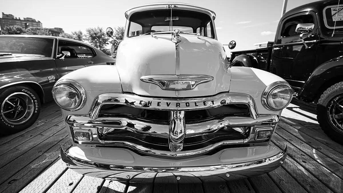 A black and white photograph primarily featuring the front of a classic Chevrolet 3100 pickup truck parked on a wooden surface.