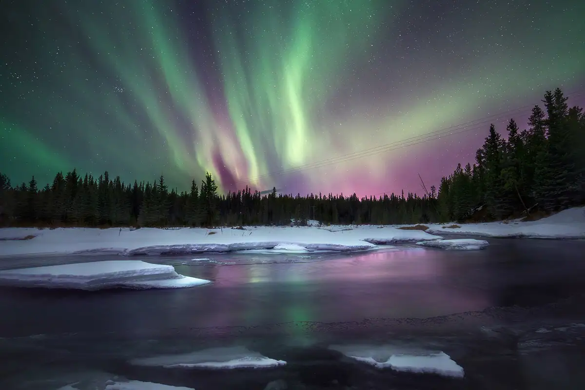Purple and green northern lights reflect in a calm body of water surrounded by snow.