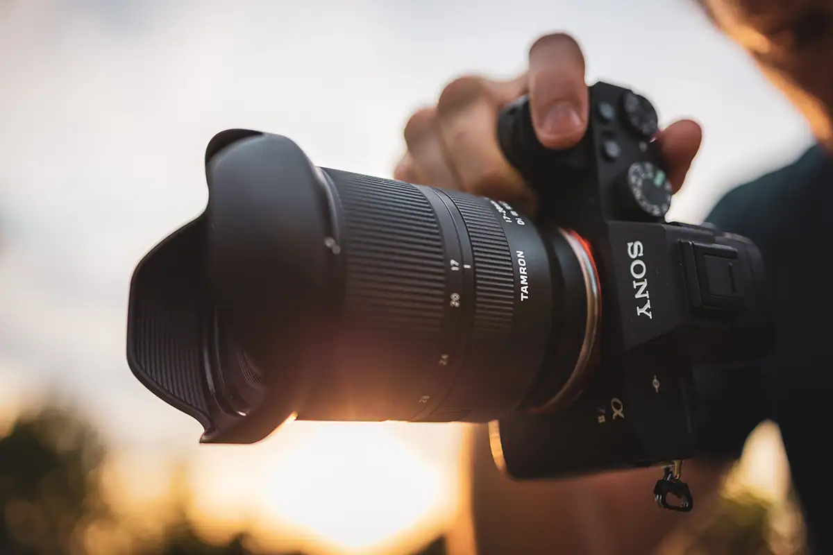 Close-up de um fotógrafo segurando uma câmera Sony equipada com uma lente Tamron, capturada sob luz solar quente com reflexo de lente - ilustrando a qualidade e a sensação de uma atualização de lente profissional.