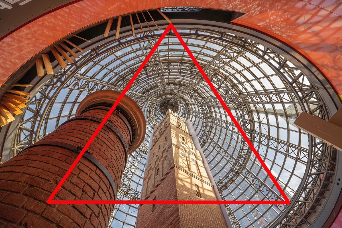 Upward view of a brick tower framed by a glass dome ceiling with a red triangle overlay, demonstrating triangular composition.
