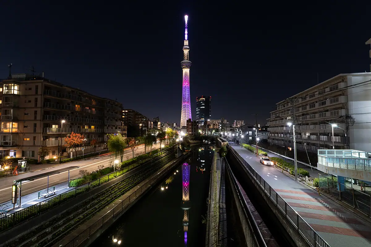 Tokyo Skytree iluminado por la noche con luces y reflejos de la ciudad, capturado en formato de imagen RAW para obtener detalles con poca luz y alto contraste.