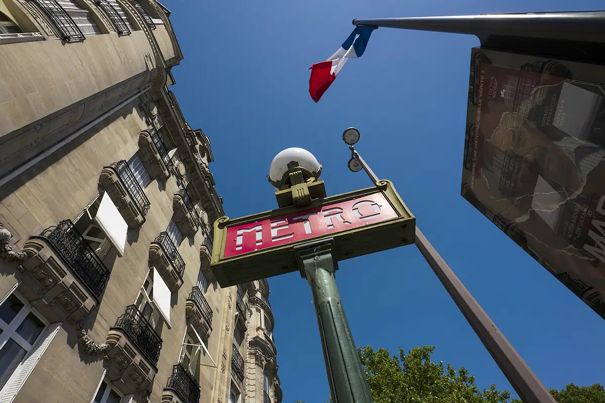 Vue en contre-plongée d'un panneau "METRO" rouge et vert monté sur un grand poteau vert, dans un ciel bleu clair. On aperçoit un drapeau français flottant sur un mât au-dessus de l'enseigne et, à l'arrière-plan, un immeuble de plusieurs étages avec des balcons.