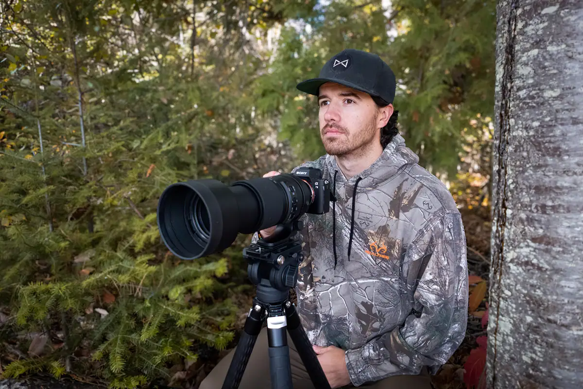 Un fotógrafo de fauna salvaje vestido con camuflaje y gorra se sienta tranquilamente en el bosque con una cámara Sony y un teleobjetivo montados en un trípode, listo para el seguimiento de animales en fotografía de fauna salvaje.