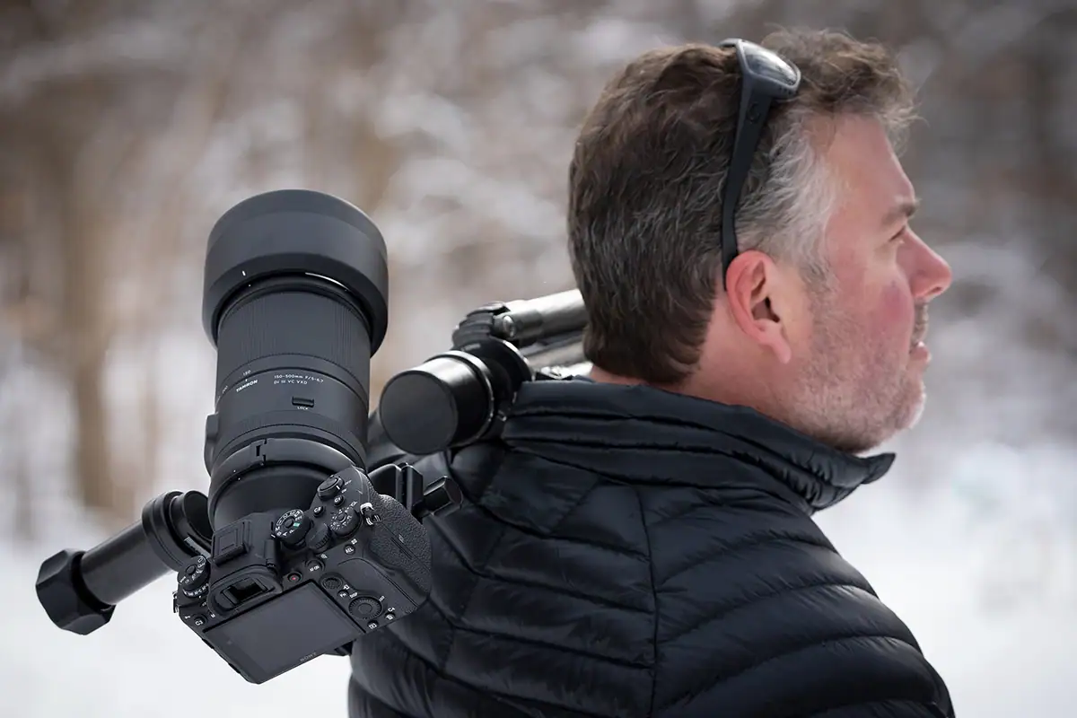 A wildlife photographer carrying a camera with a long telephoto lens and tripod over his shoulder in a snowy forest, preparing to capture shots for winter wildlife photography tips.