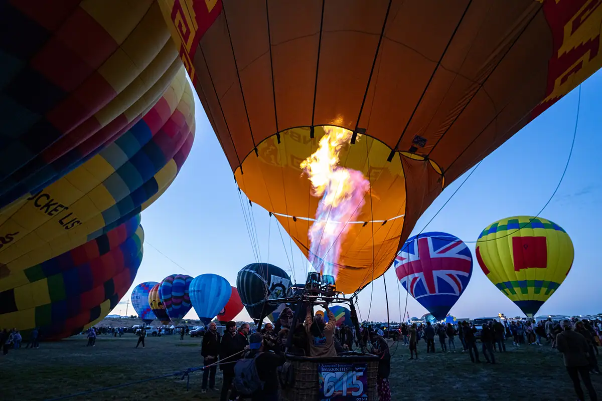 Hot air balloons inflating at blue hour with flames lighting one balloon.