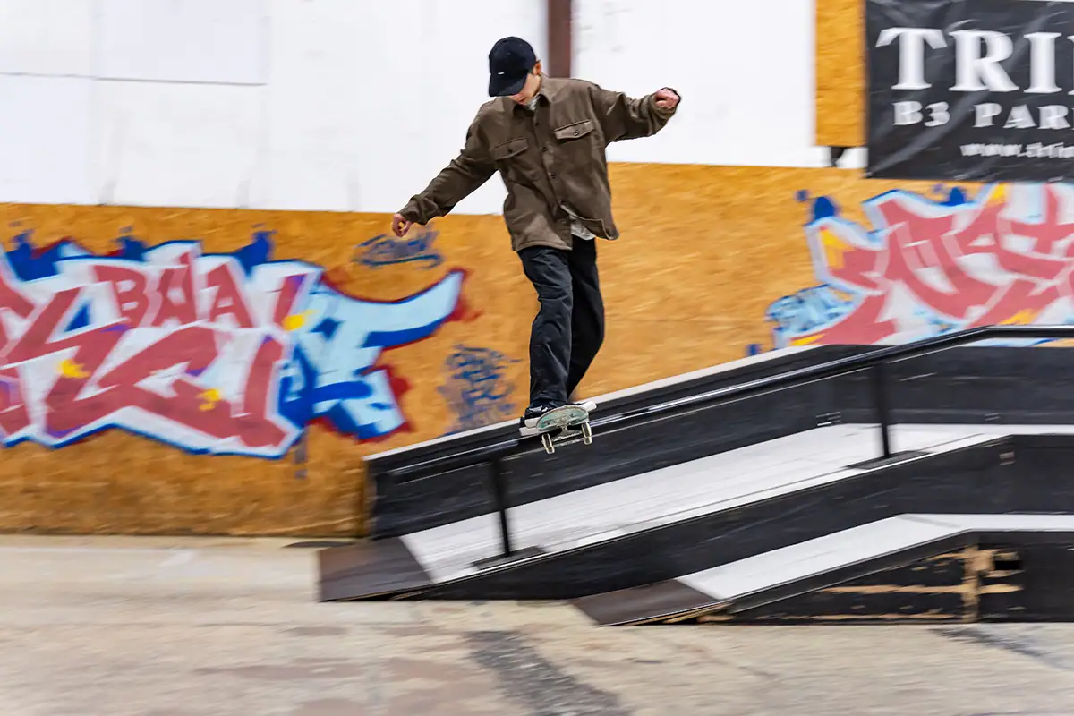 Fotografía panorámica de un patinador haciendo un grind en una barandilla en un parque de patinaje cubierto, con un fondo desenfocado que enfatiza la velocidad y el equilibrio.