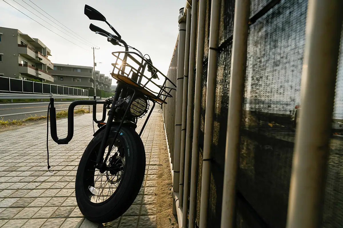 A bicycle with a front basket leans against a fence on a quiet city sidewalk at sunset, illustrating urban street photography with light and shadow.