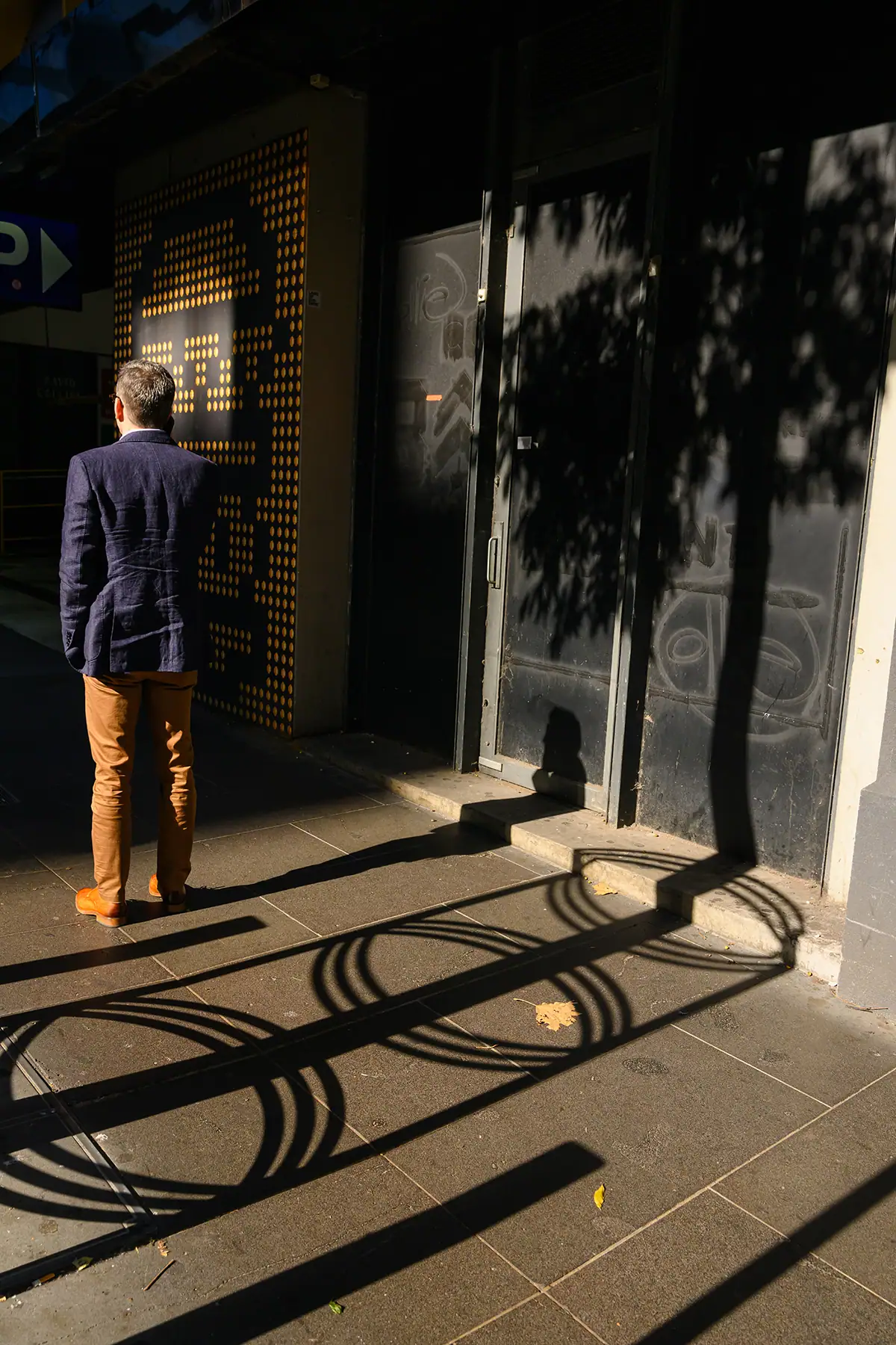 A man in a blazer stands on a sunlit city sidewalk, his long shadow stretching toward a building wall, illustrating street photography that plays with light and geometry.