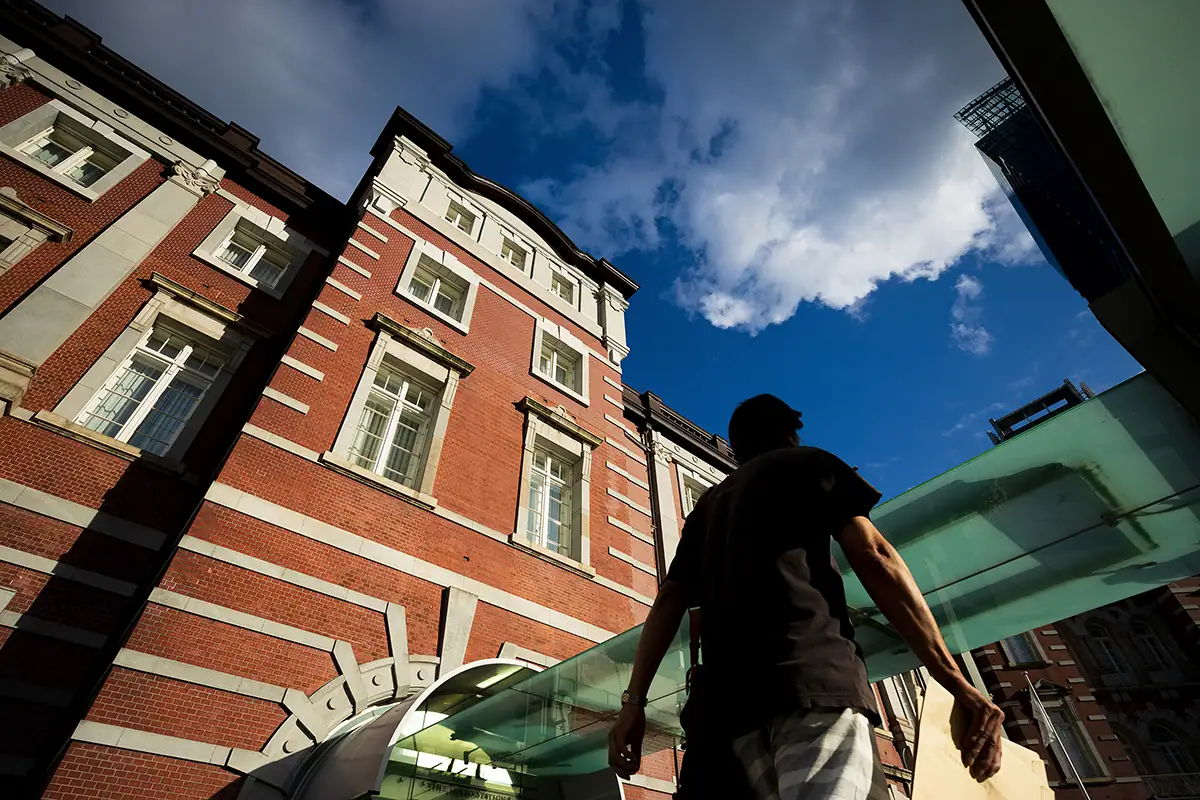 A person walks near a tall red-brick building under a bright blue sky with clouds, illustrating street photography that emphasizes architecture and perspective.