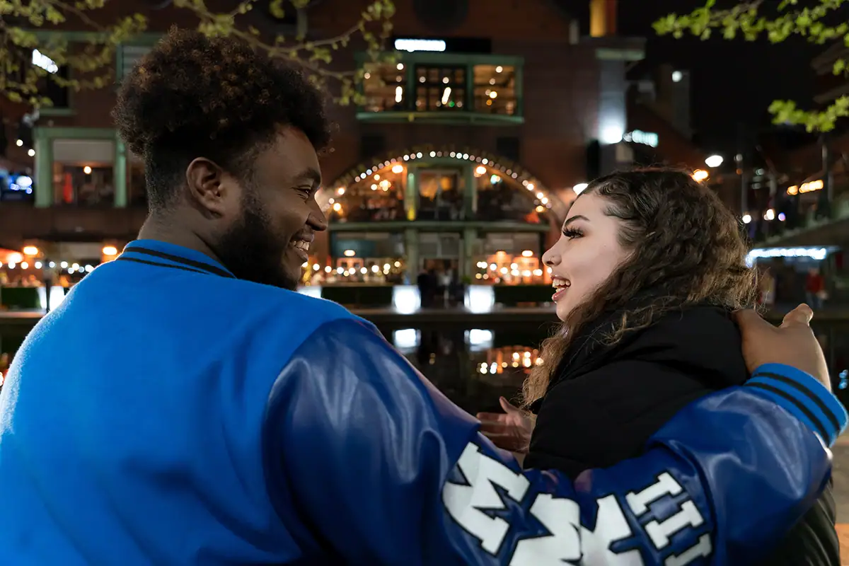 A man and woman laugh together at night near a canal lined with lights and restaurants, showing the warmth of street photography in an urban nightlife setting.