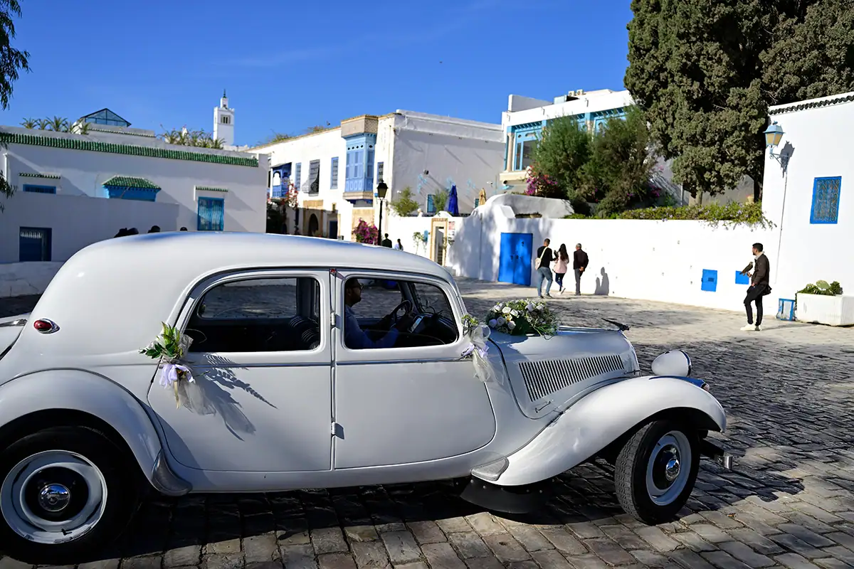 A vintage white wedding car decorated with flowers drives through a bright Mediterranean street, an example of street photography capturing culture and celebration.