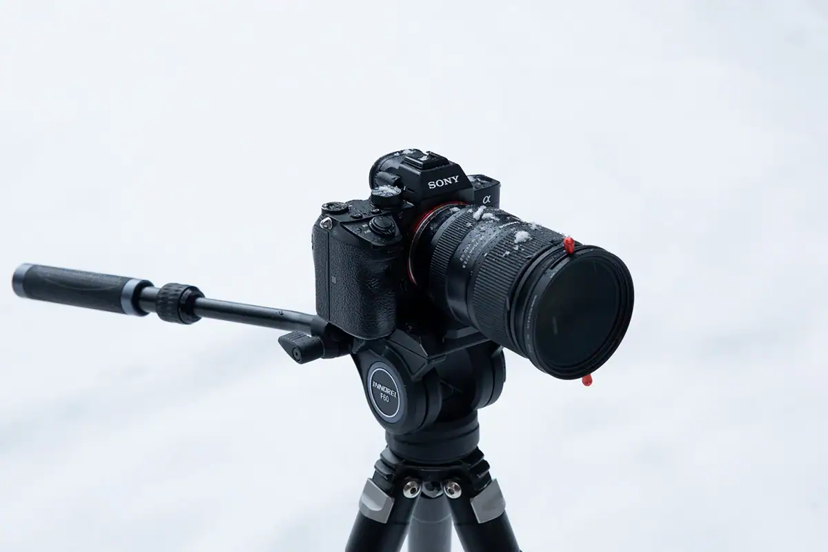 Appareil photo sans miroir Sony monté sur un trépied équipé d'un objectif Tamron 28-75 G2 avec un filtre de 67 mm, avec une fine couche de neige sur l'objectif soulignant sa résistance pour les prises de vue en extérieur.