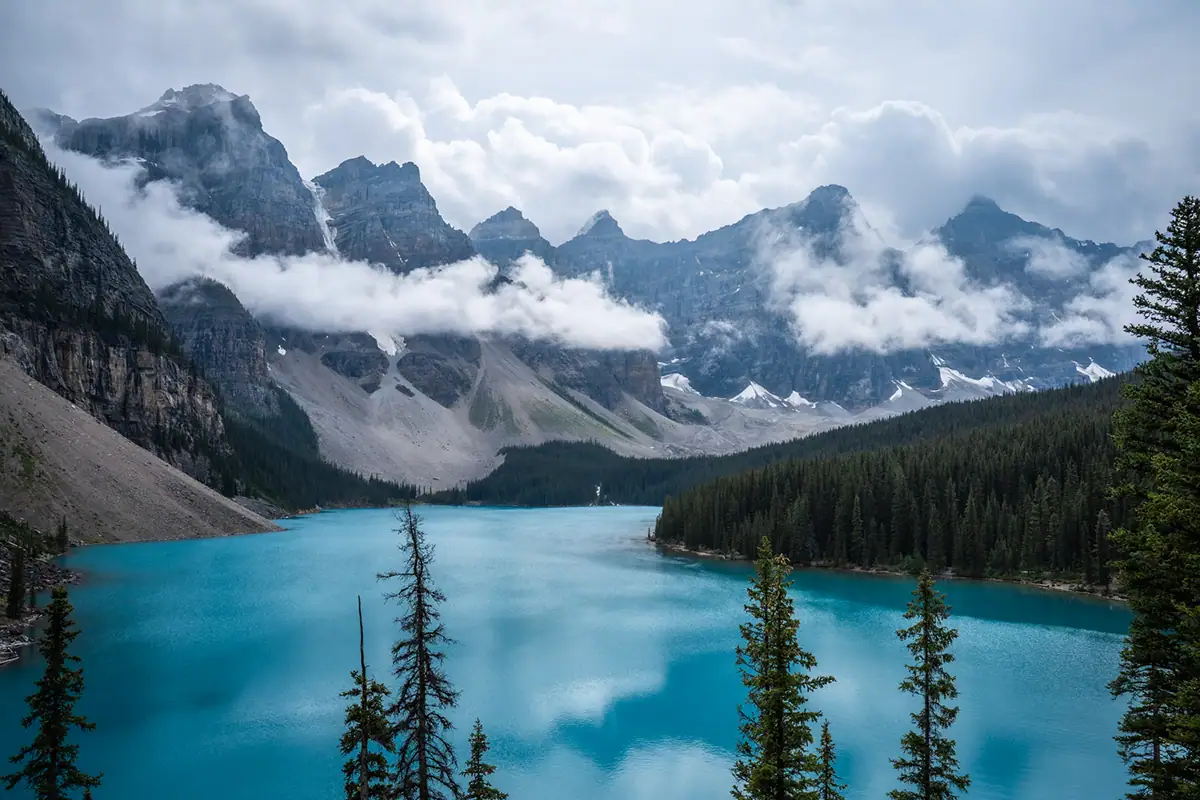 Lac turquoise éclatant entouré de forêts à feuilles persistantes et de sommets enneigés sous un ciel nuageux, capturé avec l'objectif Tamron 28-75 mm F2.8 G2 qui met en valeur les détails nets et le rendu naturel des couleurs.