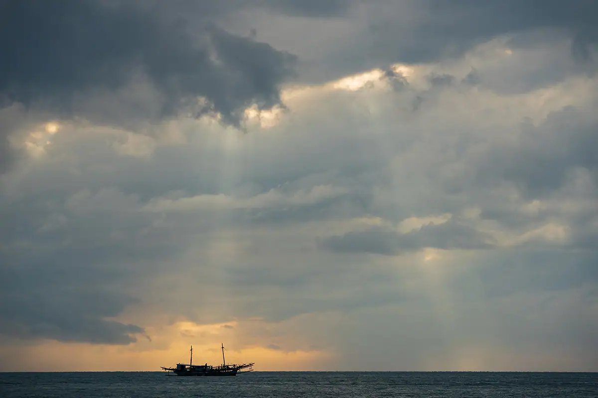 Silhouetted fishing boat on the ocean at sunset in Indonesia with dramatic clouds and sun rays, photographed during travel seascape photography with the Tamron 70-180mm F2.8 G2 lens.