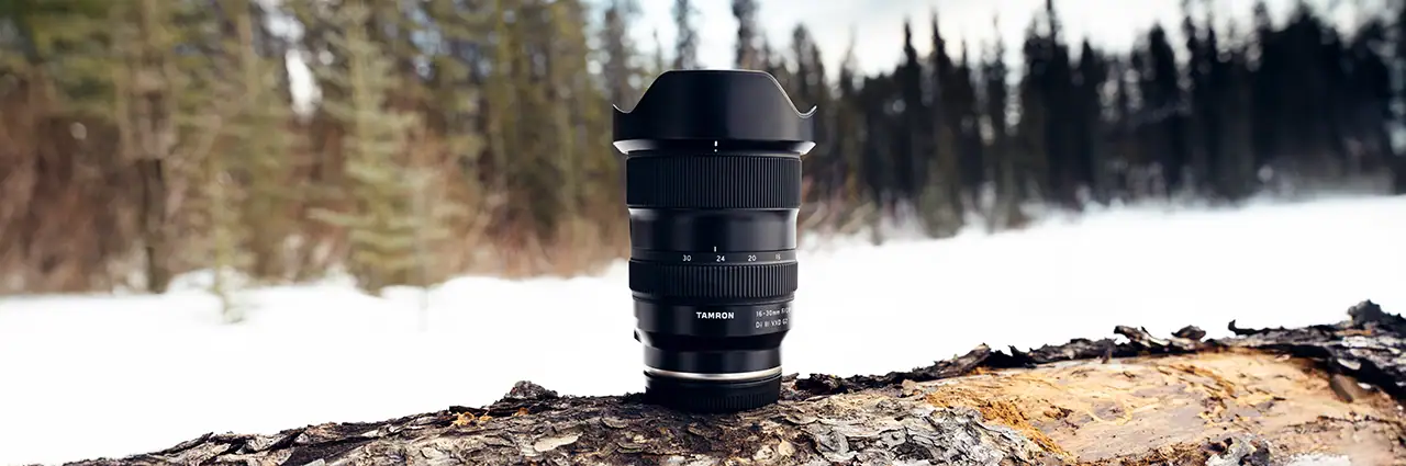 Objectif grand angle Tamron 16-30 mm F2.8 Di III VXD G2 exposé sur un tronc d'arbre dans un paysage hivernal avec une forêt de conifères floue en arrière-plan.