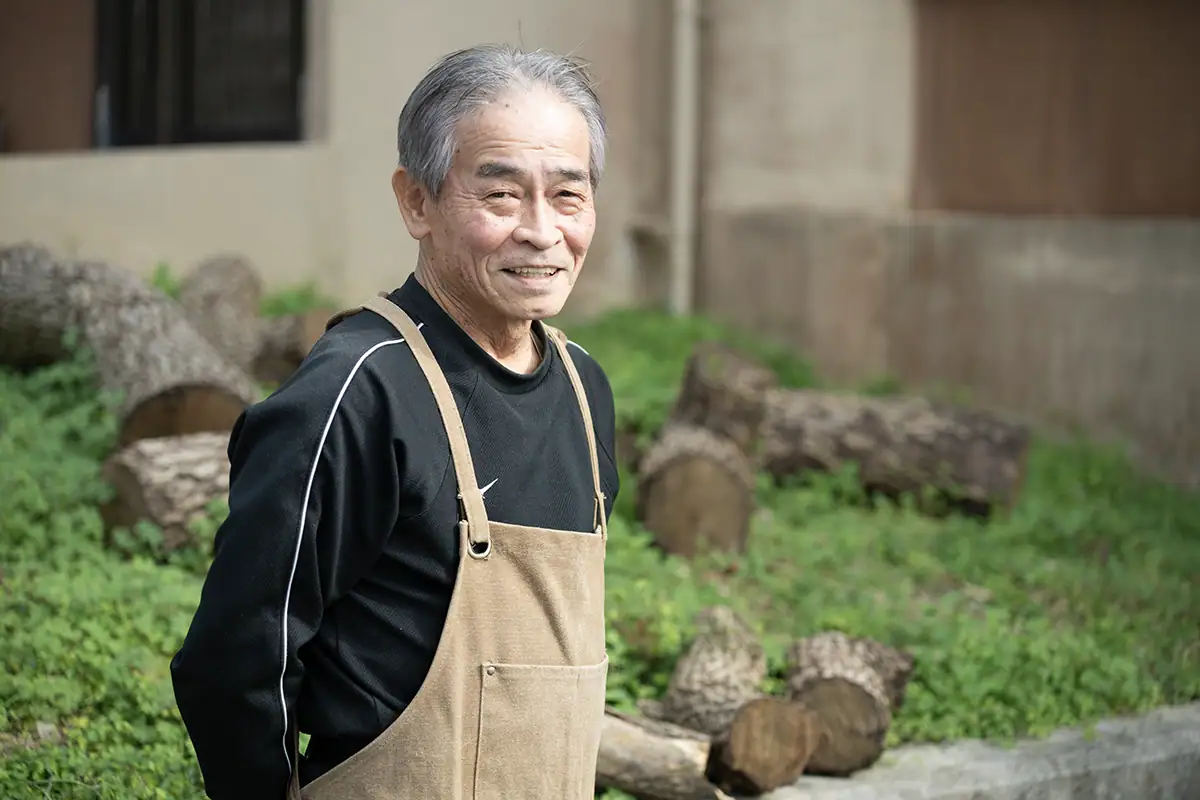 Portrait en extérieur d'un artisan d'Okinawa à côté de bois coupé, photographié avec le Tamron 28-75mm F2.8 G2 pour un documentaire de narration visuelle.