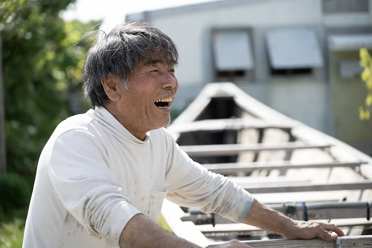 Portrait d'un constructeur de bateaux SABANI d'Okinawa riant en plein air à côté d'un cadre de coque en bois, capturé avec le Tamron 28-75mm F2.8 G2 pour un documentaire de narration visuelle.