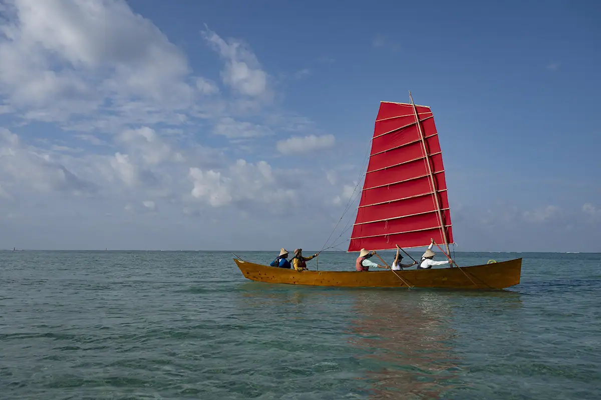 Bateau traditionnel SABANI d'Okinawa avec une voile rouge vif glissant sur une eau bleue claire, photographié avec le Tamron 28-75mm F2.8 G2 pour un documentaire culturel.