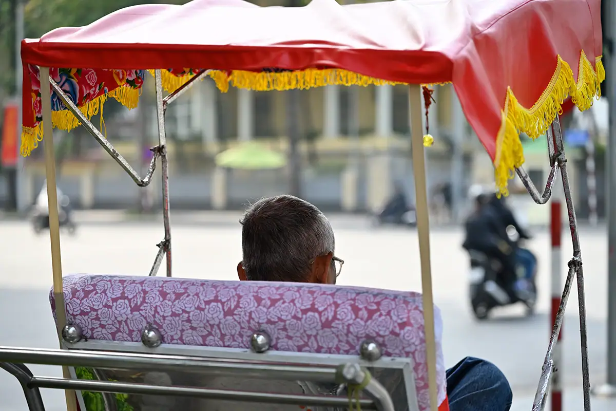 Vista trasera de un conductor de ciclo sentado bajo un toldo rojo en una calle luminosa de Vietnam, fotografiado con el Tamron 28-75 mm F2.8 G2.