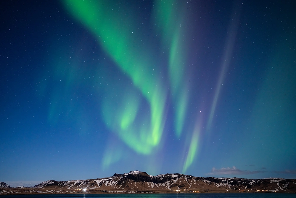 Aurora boreal fotografiada con el objetivo Tamron 35-150 mm F2-2.8, que muestra vibrantes patrones aurorales verdes sobre una cordillera nevada bajo un cielo nocturno despejado.