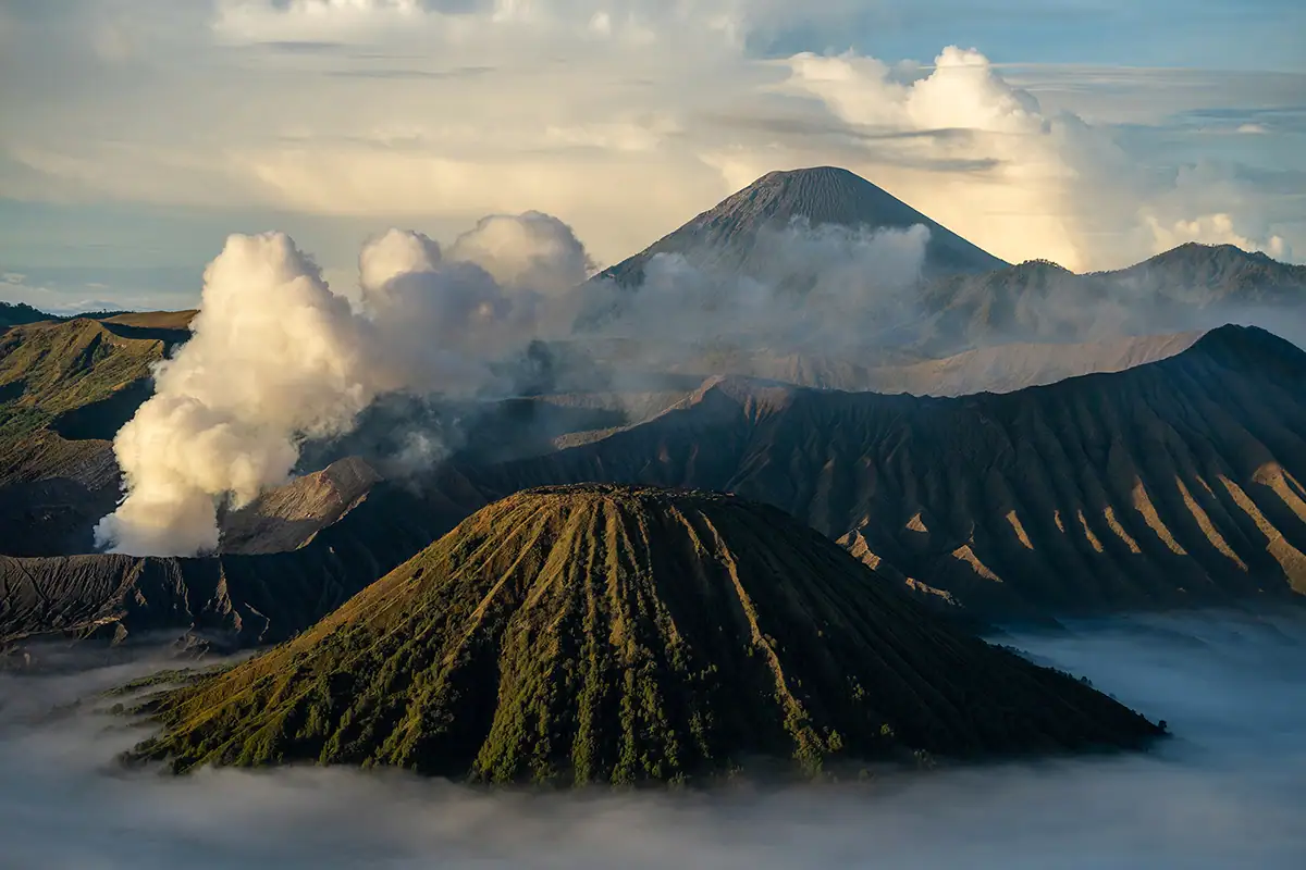Wide landscape view of volcanic peaks and drifting clouds in Indonesia captured at sunrise, showing dramatic textures, atmospheric depth, and long-range telephoto detail.