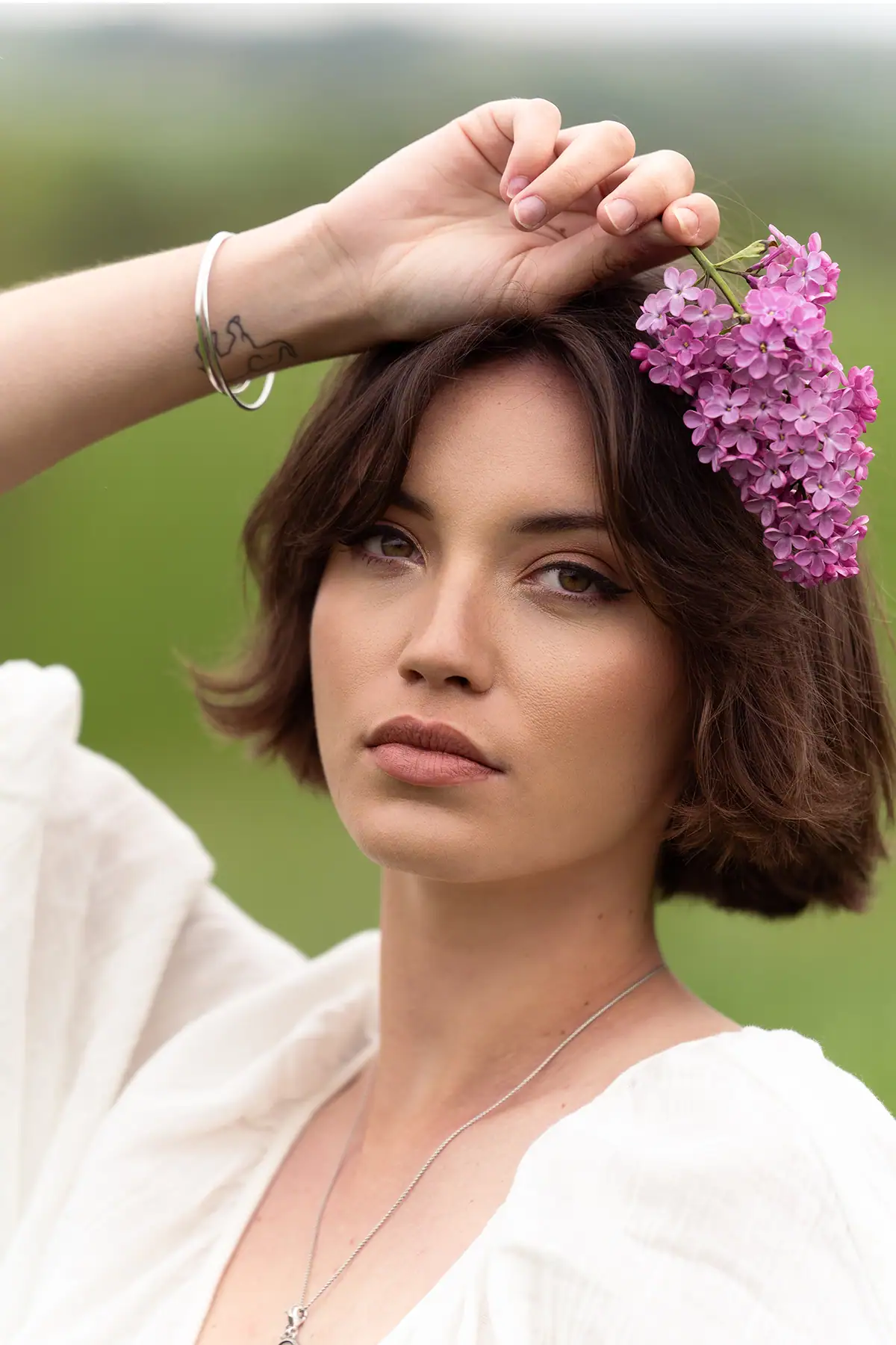 Portrait of a woman outdoors holding lilac flowers above her head, photographed with a telephoto lens to showcase soft background blur, natural skin tones, and detailed eye focus.