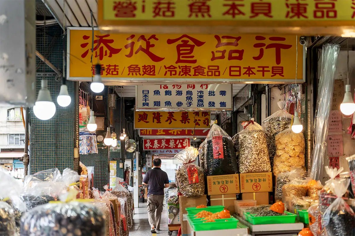 Un voyageur se promenant dans un marché en plein air animé, bordé de lumières suspendues, de produits séchés et d'enseignes colorées, capturé pour mettre en valeur le récit de voyage au quotidien.