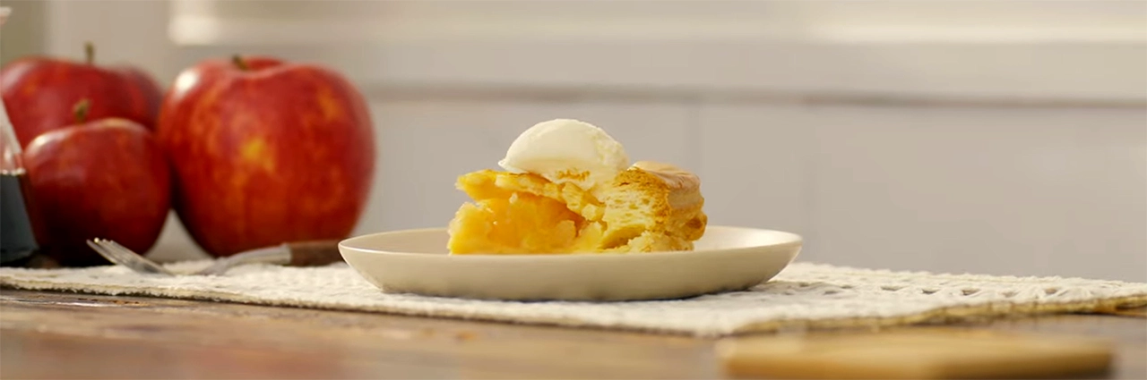 Image de héros d'une tranche de tarte aux pommes garnie de crème glacée sur une assiette, avec des pommes en arrière-plan et un doux bokeh cinématique.