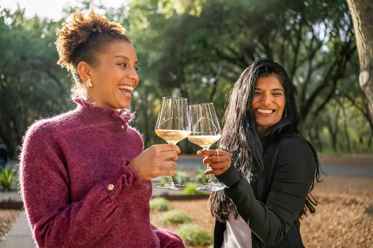 Deux femmes souriant et trinquant avec des verres de vin blanc en plein air, dans une lumière naturelle chaleureuse, entourées d'arbres.
