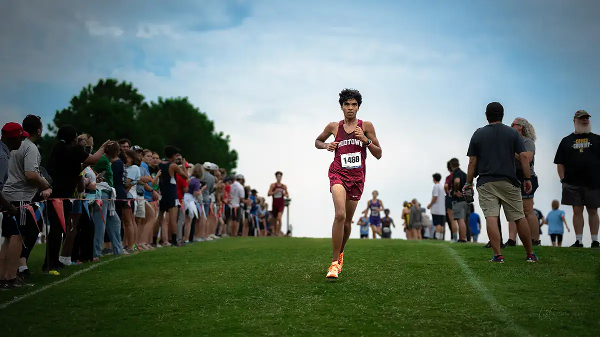A young man running cross-country track with a crowd watching