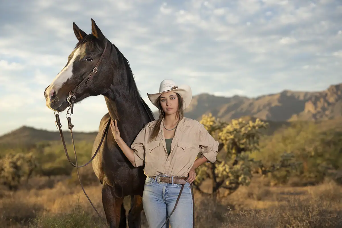 Cowgirl standing beside a horse in a desert landscape with mountains in the background.