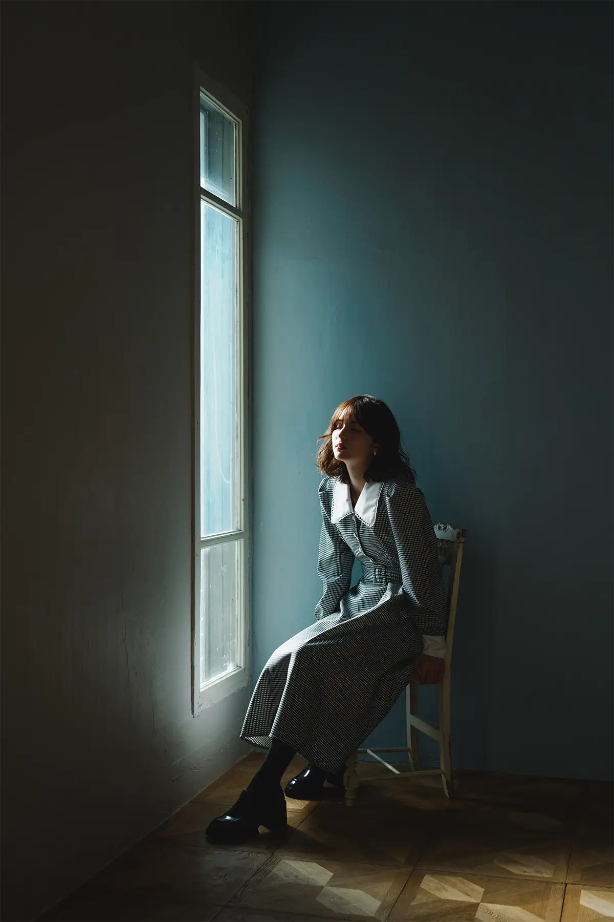 Woman seated beside a window in soft directional light with deep shadow and strong negative space.
