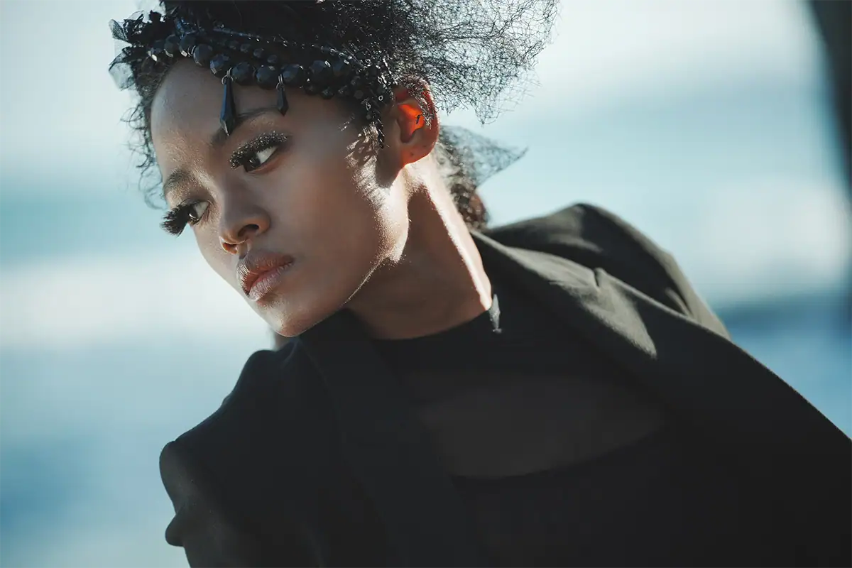 Fashion portrait of a model in black styling with dramatic makeup against a softly blurred background.