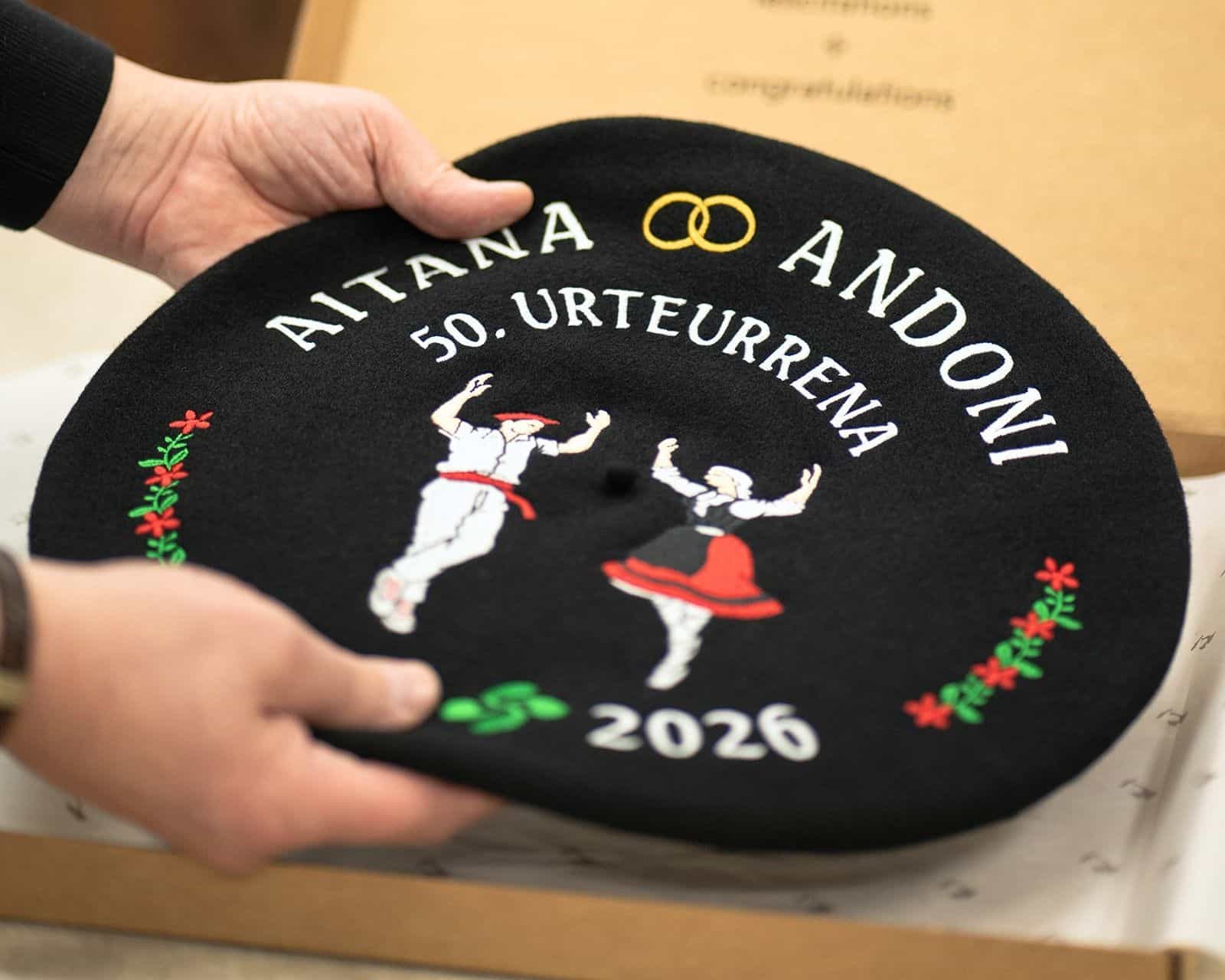 Txapela personalizada para bodas de oro con bordado de dantzaris vascos, nombres Aitana y Andoni, fecha 2026 y motivos florales, sostenida sobre caja de presentación